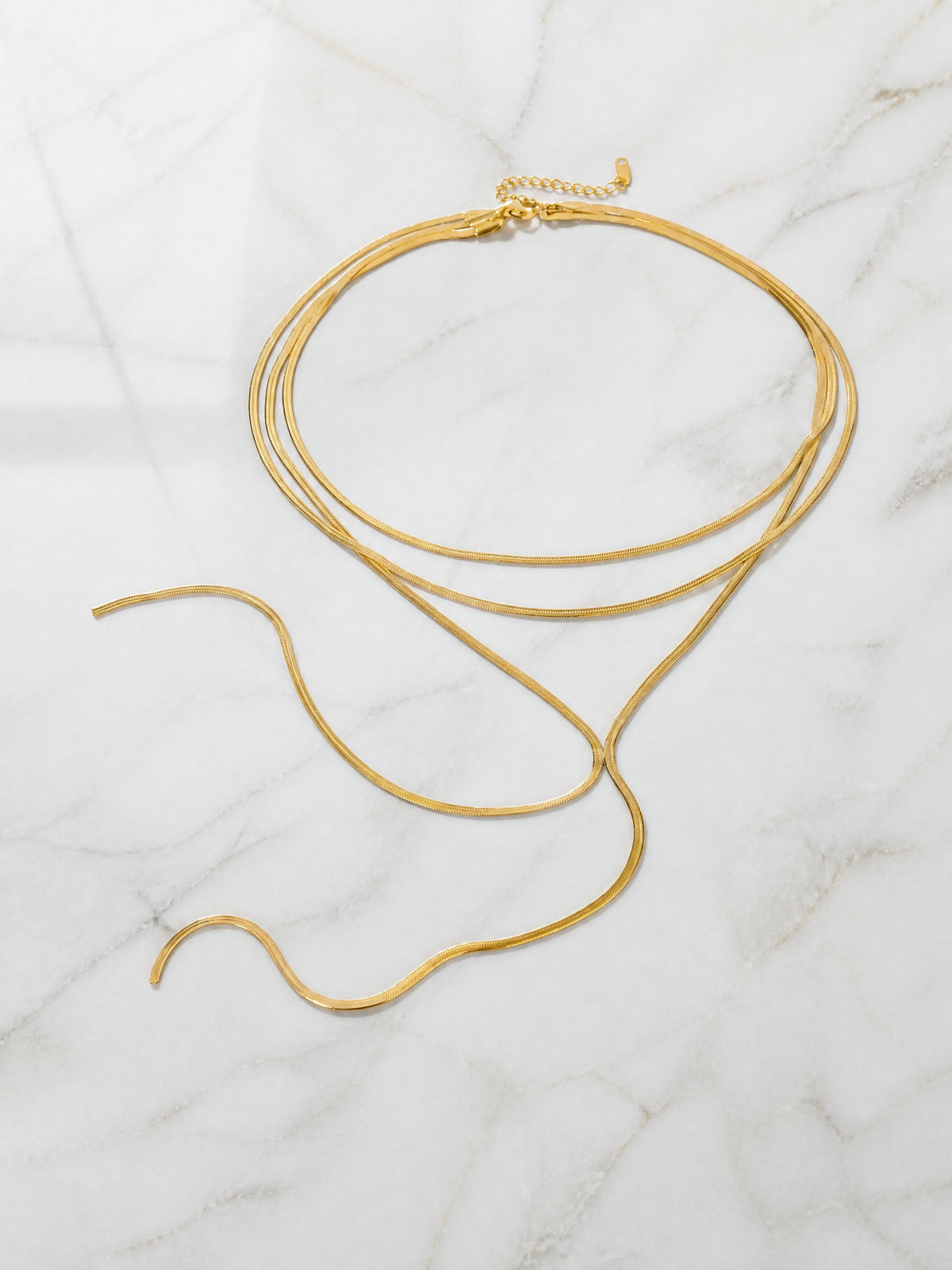 Collier Liane posé à plat sur marbre blanc – rangs de maille plate dorée, cravate en cascade à deux brins et chaînette d'extension
