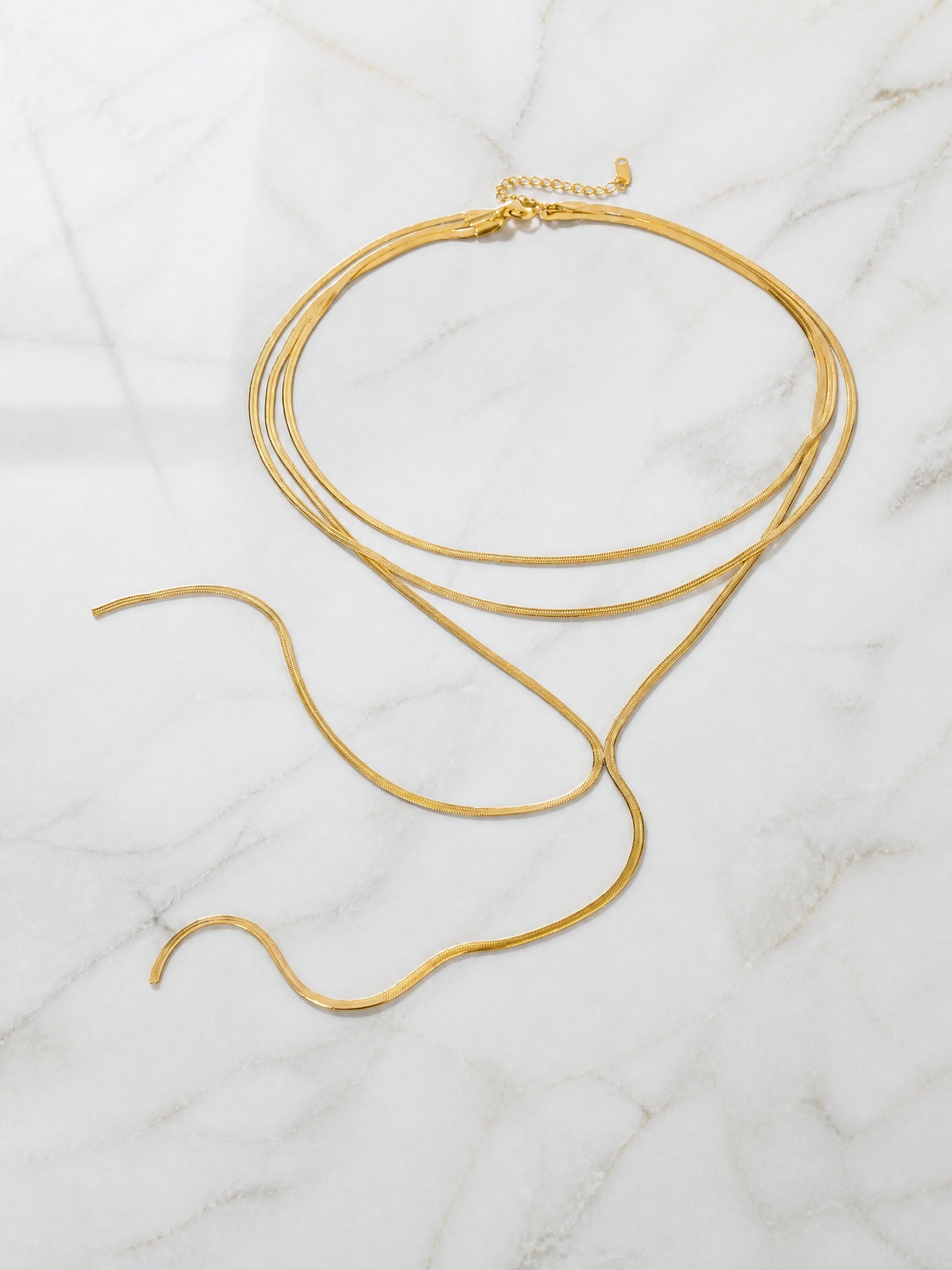Collier Liane posé à plat sur marbre blanc – rangs de maille plate dorée, cravate en cascade à deux brins et chaînette d'extension