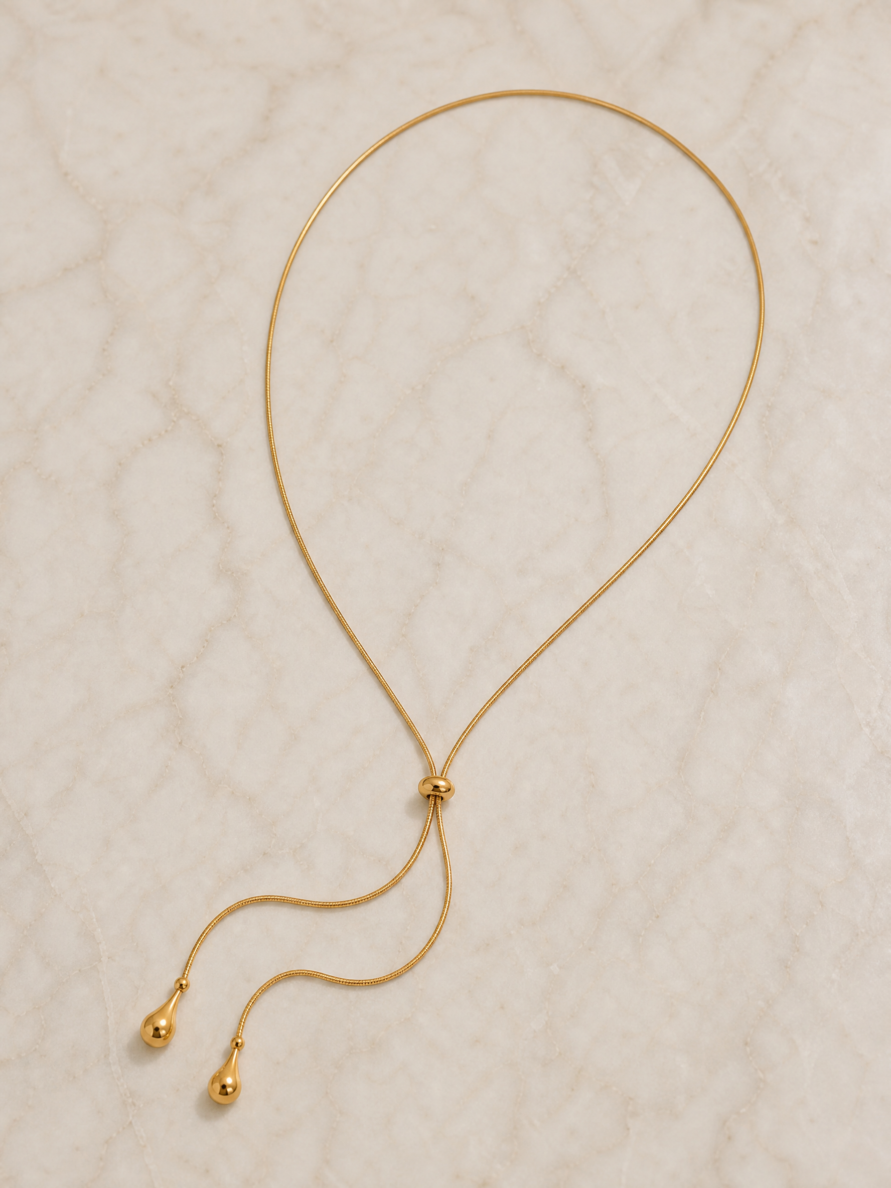 Collier Thaïs à plat sur marbre crème : maille serpent, nœud coulissant et deux gouttes dorées